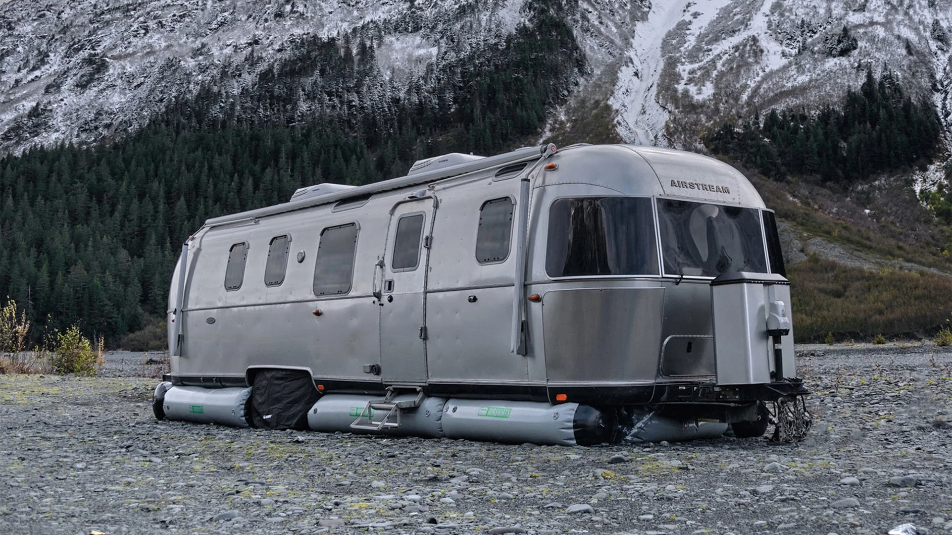 A modern, silver Airstream trailer on inflatable pontoons parked on rocky terrain with snow-capped mountains and a dense forest in the background.