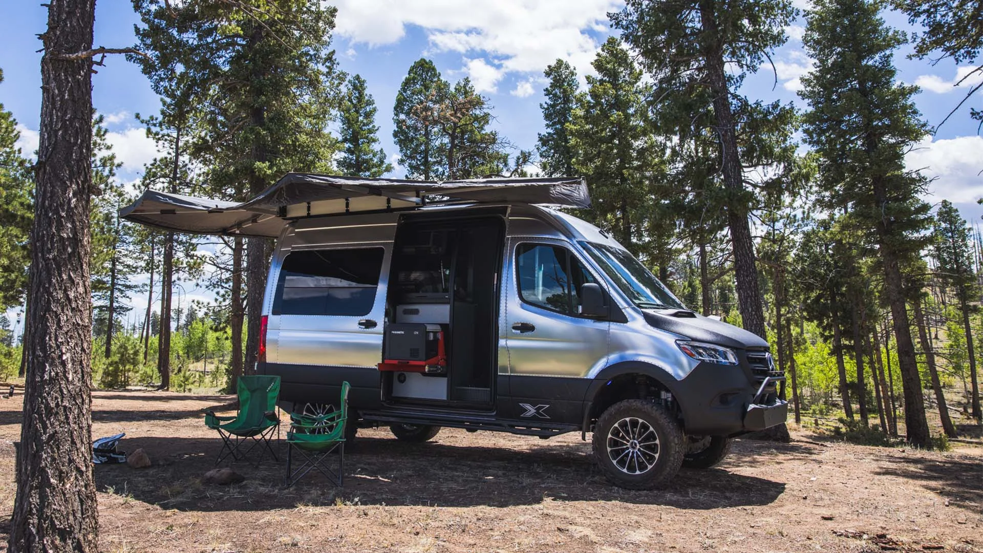 Camper van parked in a forest clearing with an awning extended, surrounded by tall pine trees under a clear blue sky.