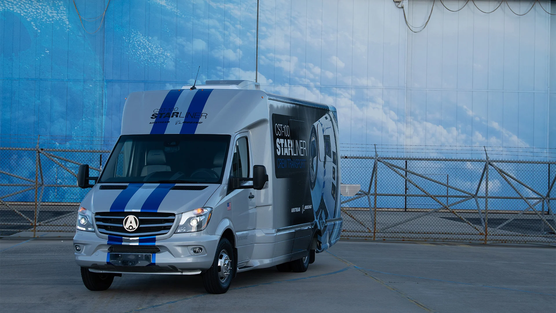 Silver Starliner van with blue accents parked in front of a large blue and white wall, featuring clouds.