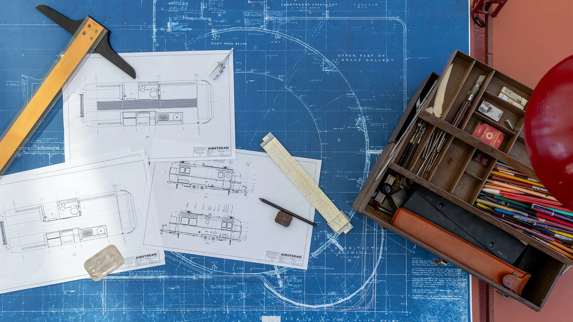 Blueprints, drafting tools, and an open toolbox with pencils and instruments are arranged on a blue drafting table.
