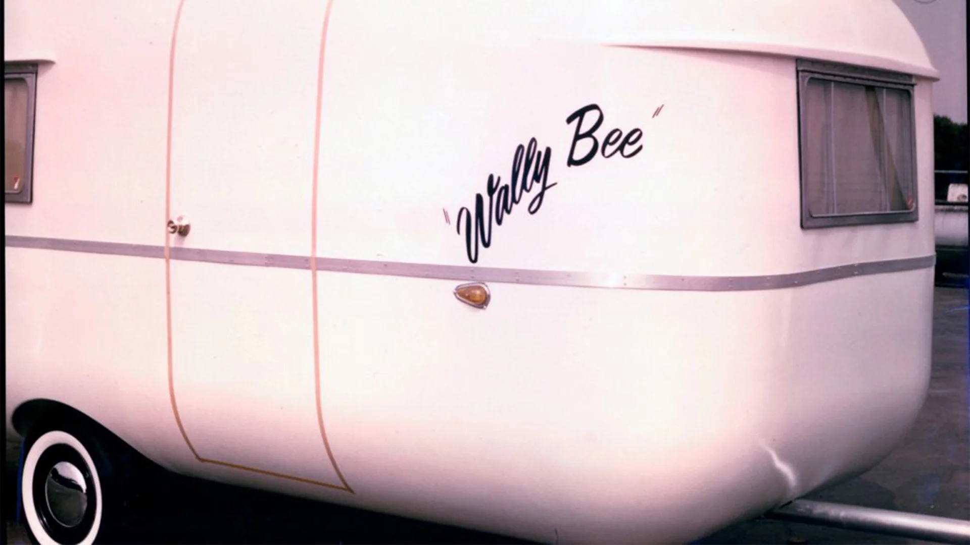 Side view of a vintage white trailer with "Wally Bee" written on it. It has a rounded shape and a small round light near the wheel.