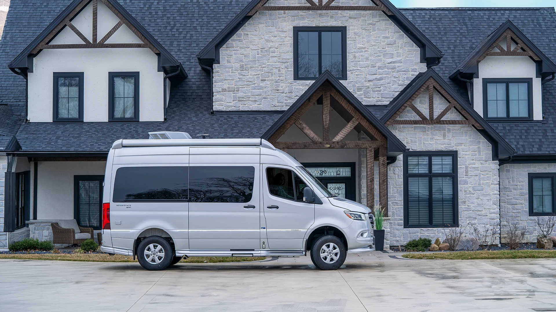 A silver van parked in front of a large, modern house with a stone exterior and dark trim, featuring gabled roofs and multiple windows.