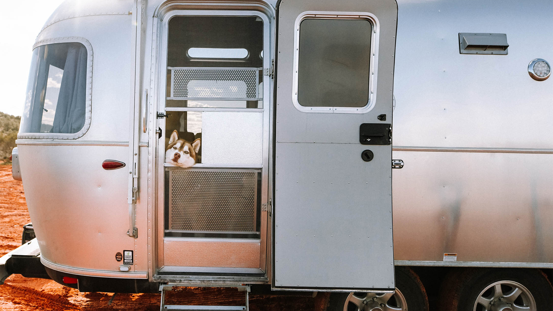 A husky peeks out from behind a screen door of a silver trailer against a red dirt landscape.