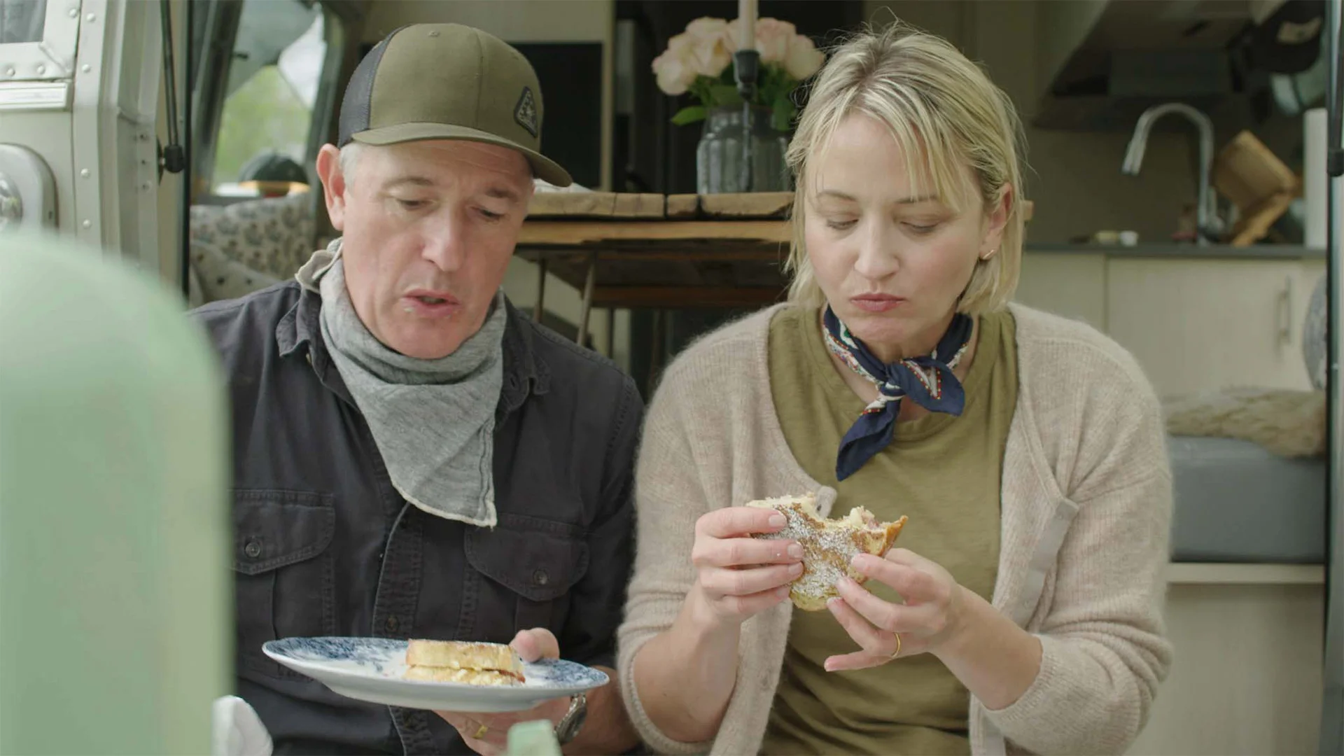A man and woman sit inside a camper van, eating sandwiches. The table behind them holds flowers and other items. They appear focused on their meal.