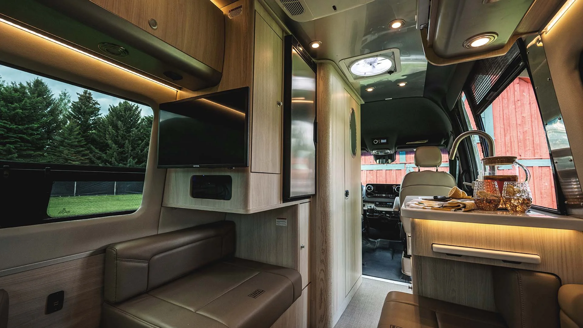 Interior of a modern camper van with wood finish, featuring seating, a TV, kitchenette with a tap, glassware, and a skylight, surrounded by trees outside.