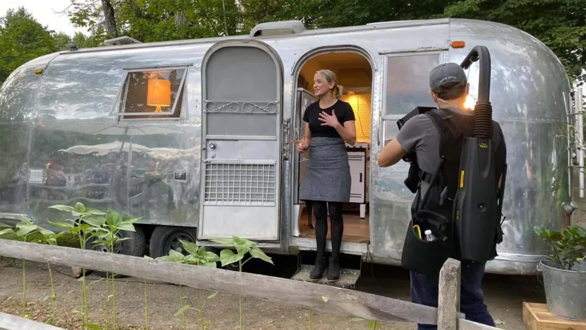 A woman stands in a shiny silver trailer's doorway, gesturing while talking to a person holding cleaning equipment. Trees and plants surround them.