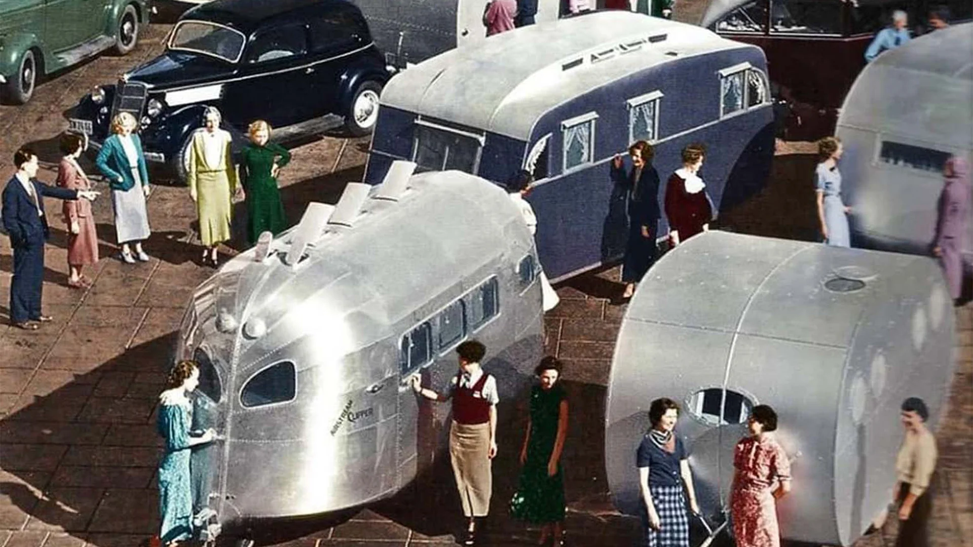 A group of people examines vintage, futuristic-style trailers and cars on display in an outdoor setting.