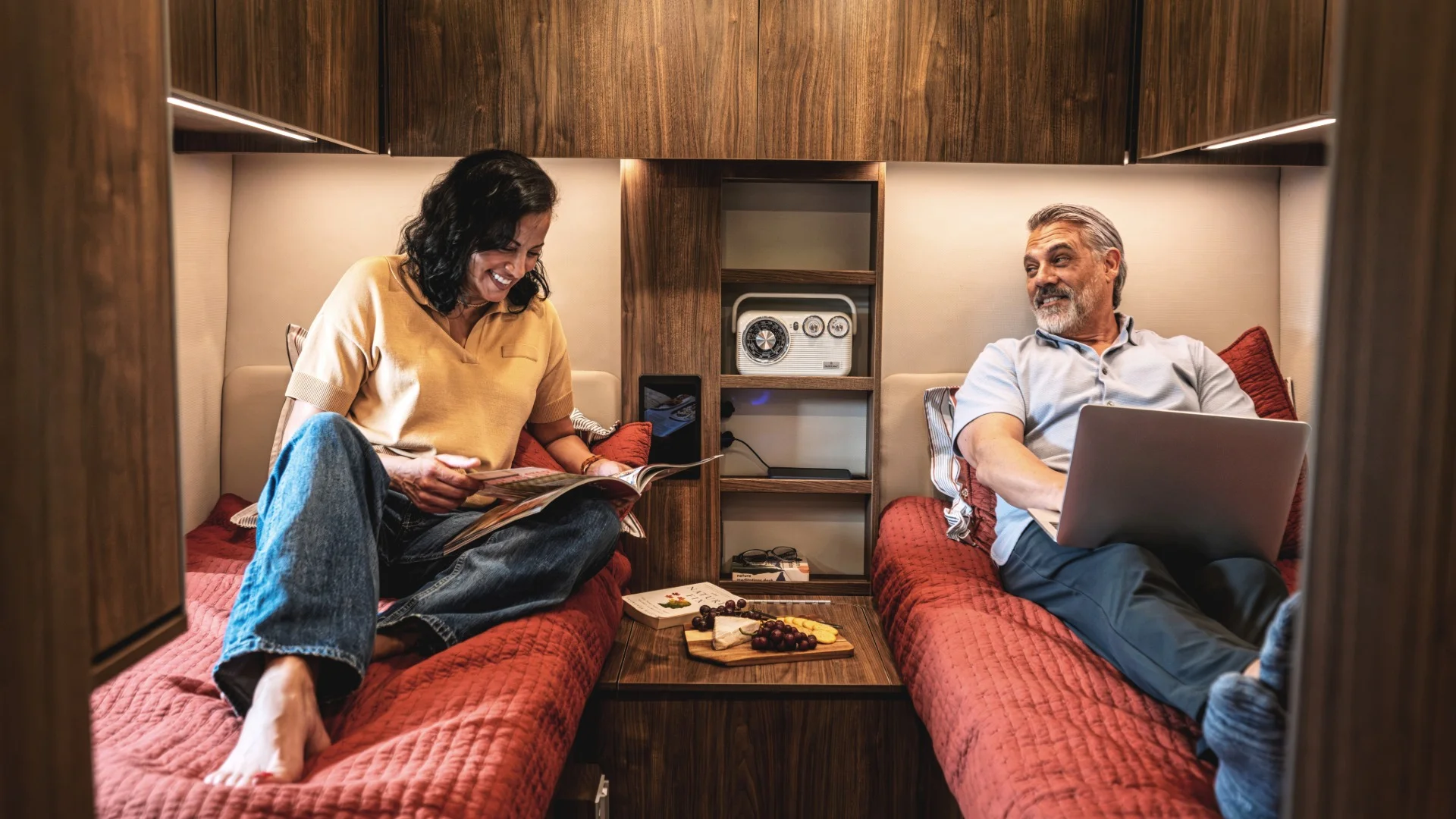 Two people relax in a camper van, each on separate beds. One reads a magazine, and the other uses a laptop. Snacks and cards are on the table between them, creating a cozy atmosphere.