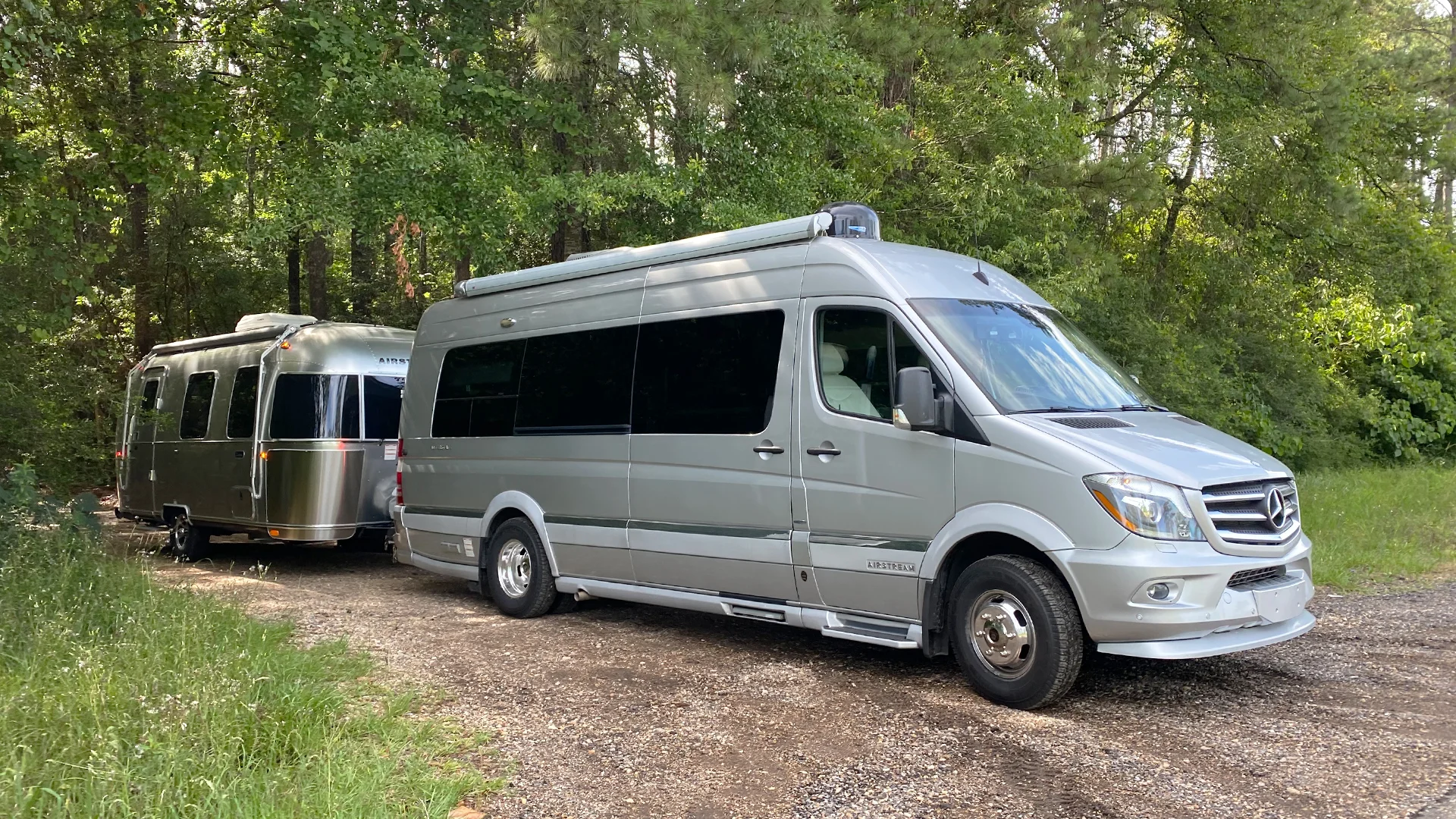 Silver camper van towing a shiny silver trailer on a gravel path, surrounded by lush green trees.