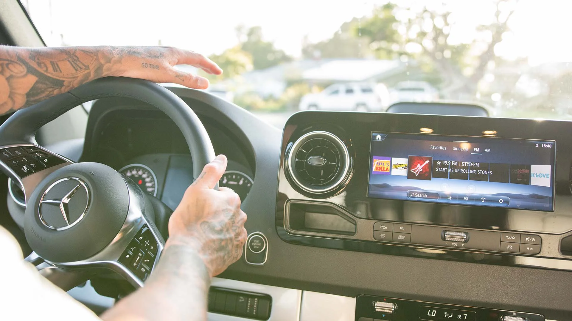 Person driving a car with a tattooed arm, steering wheel featuring a logo, and dashboard displaying music on the screen.