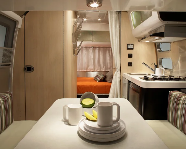 Interior of a modern RV with a dining area, kitchenette, and cozy sleeping area. Striped seats, tableware, and fruit are on the table.