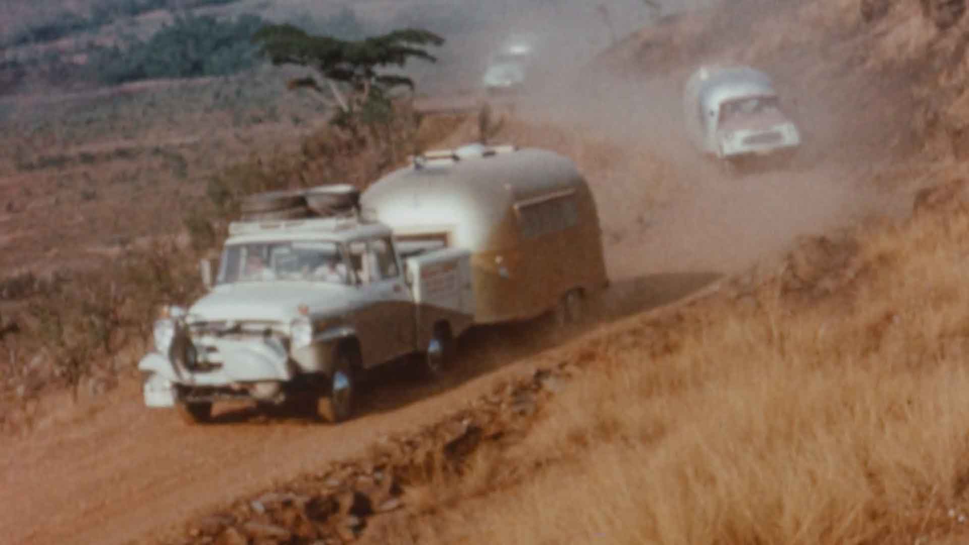 Vintage cars towing trailers on a dusty, winding road through a hilly landscape, with dry grass and sparse vegetation.