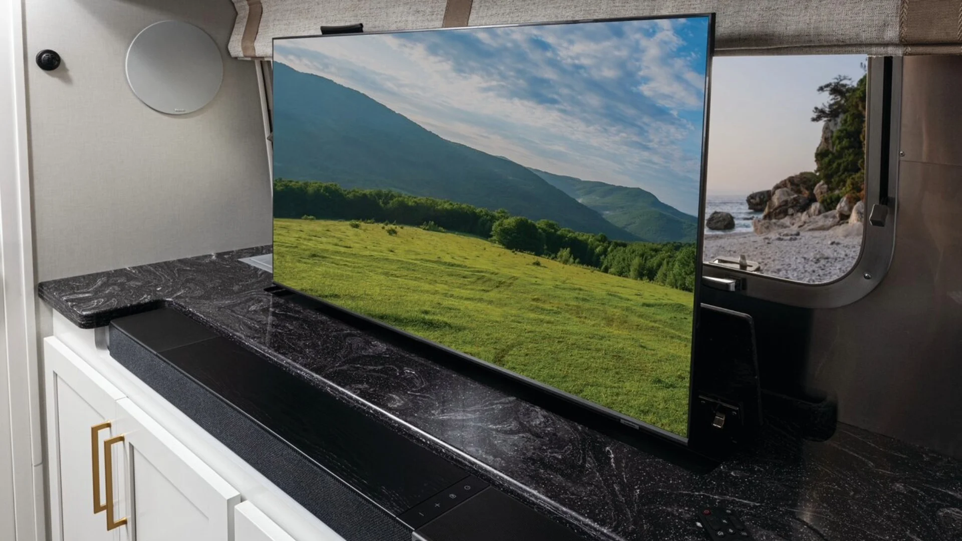 Flat-screen TV displaying a scenic landscape with mountains and green fields on a counter, near a window with a coastal view.