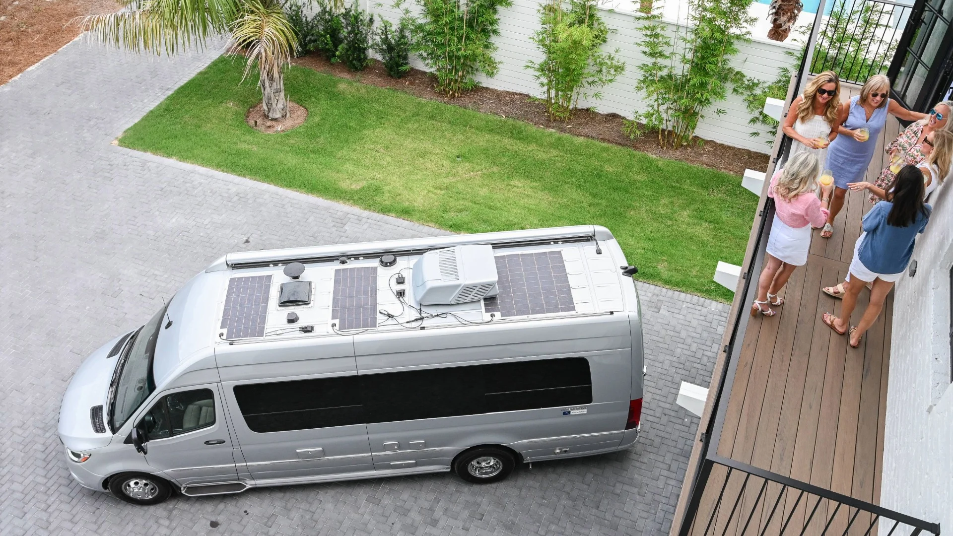A silver camper van with solar panels on the roof is parked in a driveway, while six women stand on a balcony and nearby patio, talking and smiling in a relaxed outdoor setting.