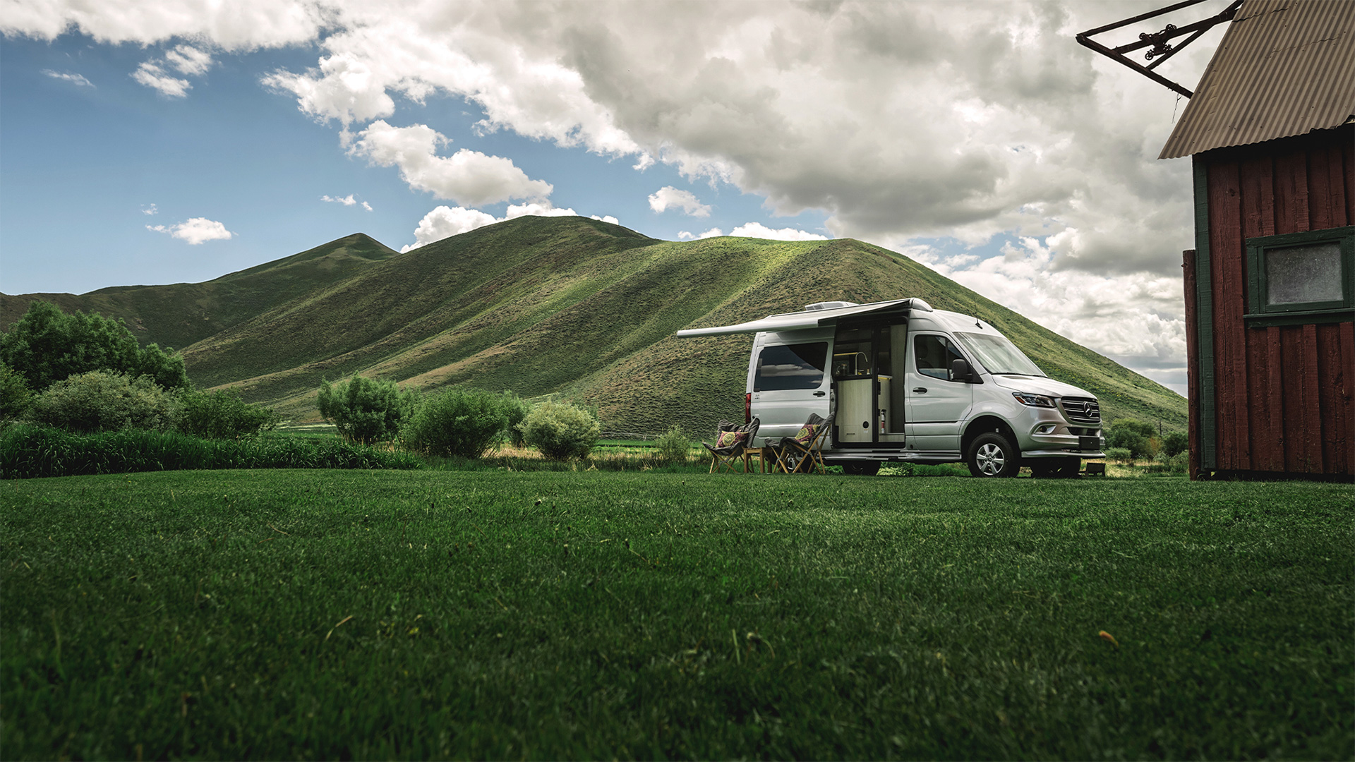 A camper van with an extended awning is parked on lush grass near a wooden building, surrounded by rolling green hills under a cloudy sky.