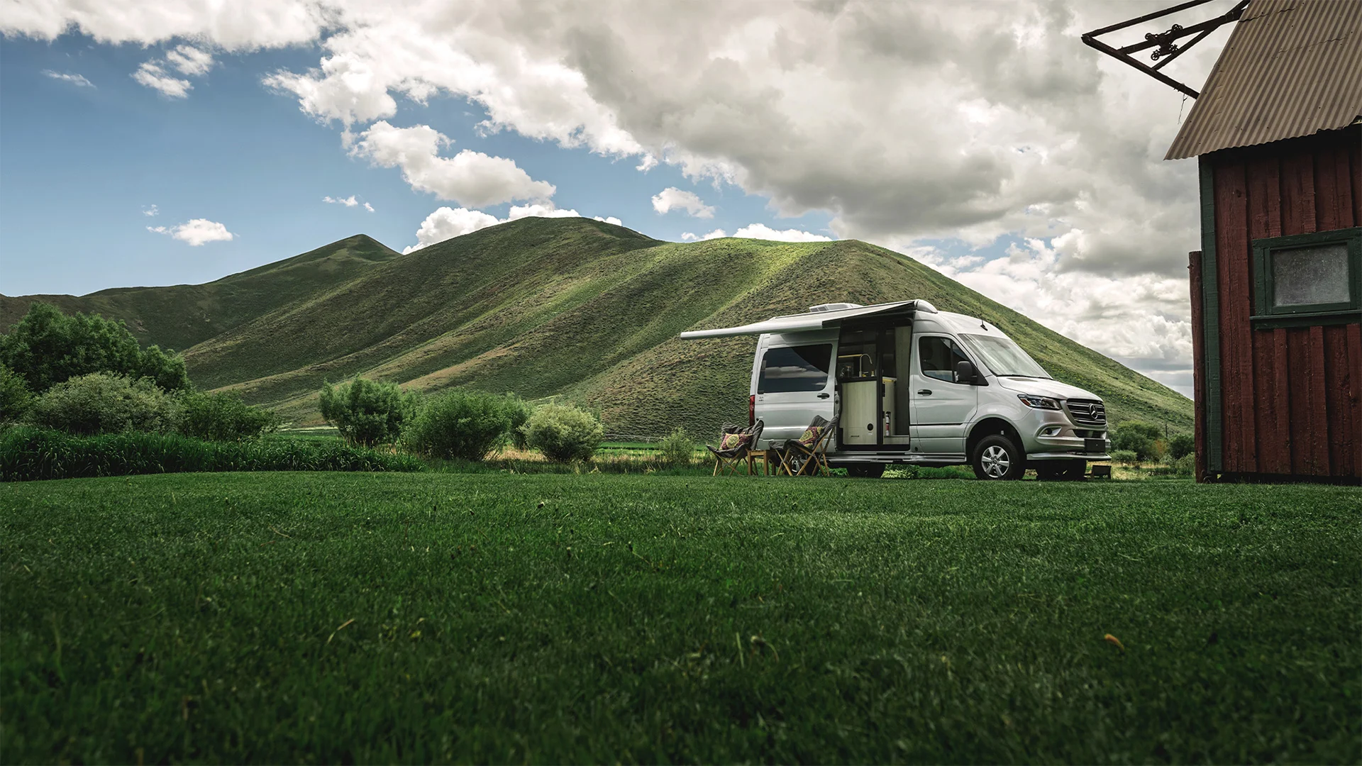 A camper van with an extended awning is parked on lush grass near a wooden building, surrounded by rolling green hills under a cloudy sky.