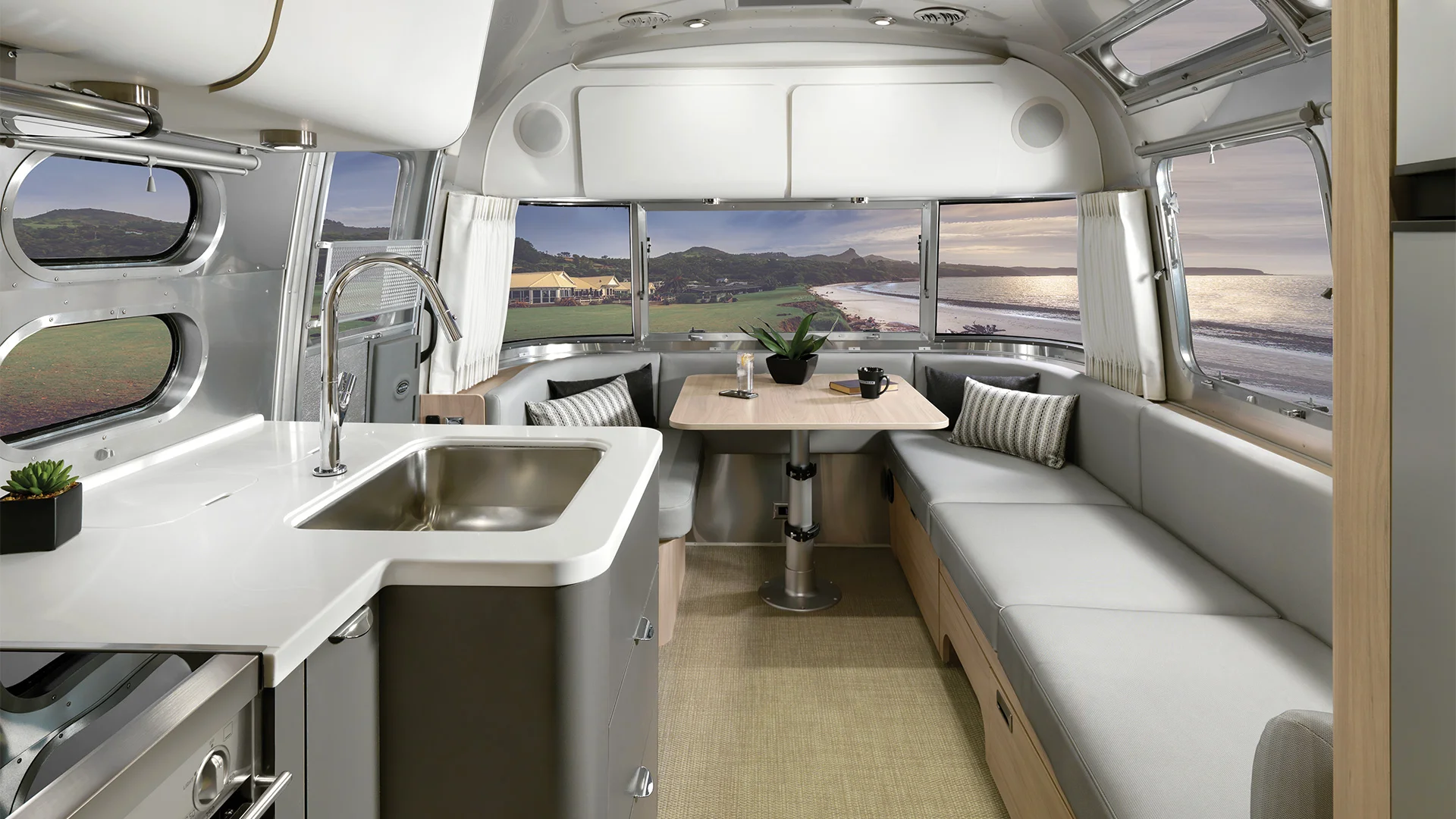 Modern camper interior with sleek kitchen, cozy dining area, panoramic windows offering a scenic coastal view at sunset.