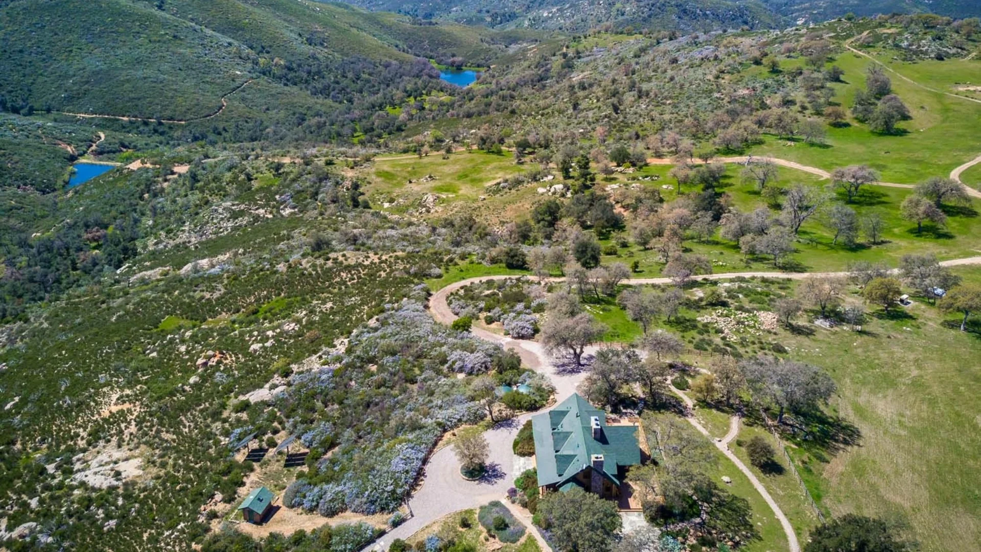 Aerial view of a lush, hilly landscape with a house at the center, surrounded by trees, greenery, and distant blue ponds.