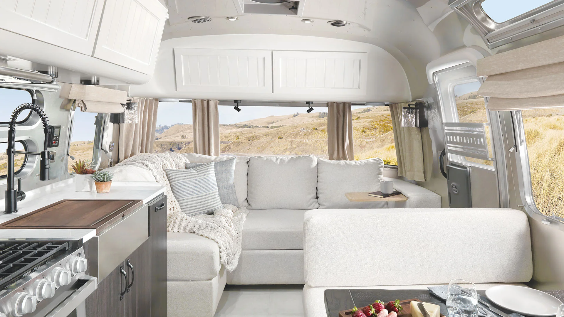 Modern RV interior with light-colored sofa, cozy pillows, and blankets next to large windows showing a scenic desert landscape. Kitchen area with stove and sink visible in the foreground. Bright and inviting atmosphere.