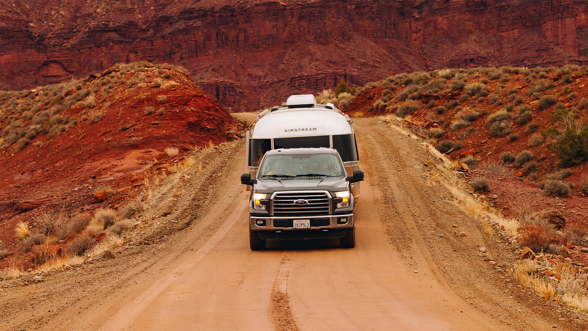 A truck towing an Airstream trailer navigates a dirt road through a rugged, red rocky desert landscape under a cloudy sky.
