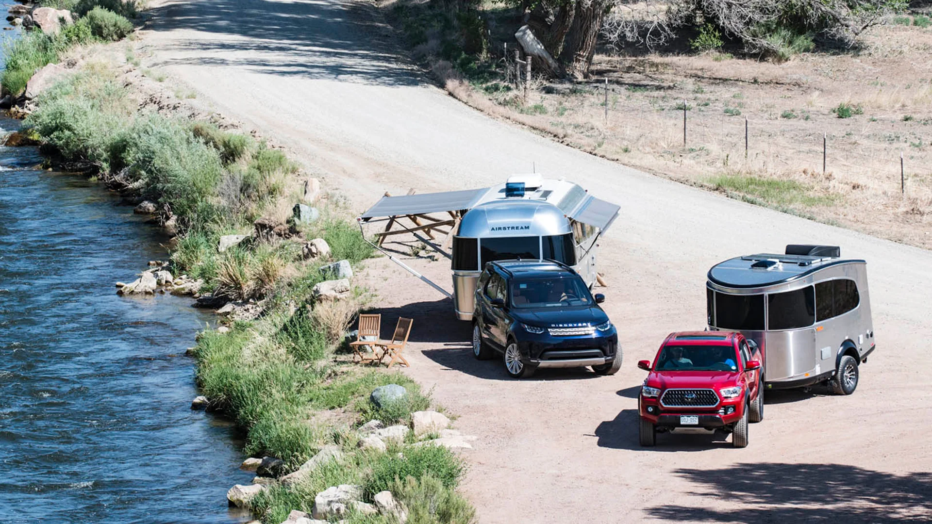 Two SUVs parked near a river, each towing a small travel trailer. One trailer has an awning extended, with chairs set up outside. The scene is surrounded by greenery and a dirt road runs alongside the river.