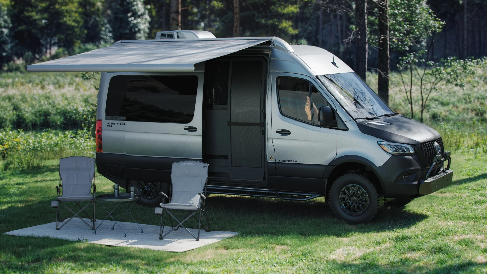 Silver camper van with extended awning, two folding chairs, and a small table set on grass in a wooded area.