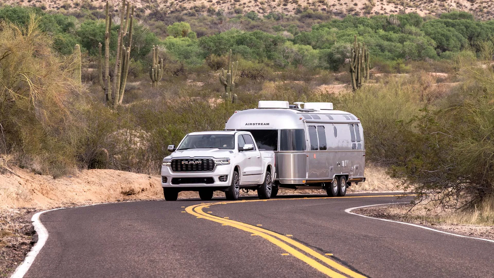 White pickup truck towing a silver Airstream trailer on a winding desert road with cacti and scrub vegetation.