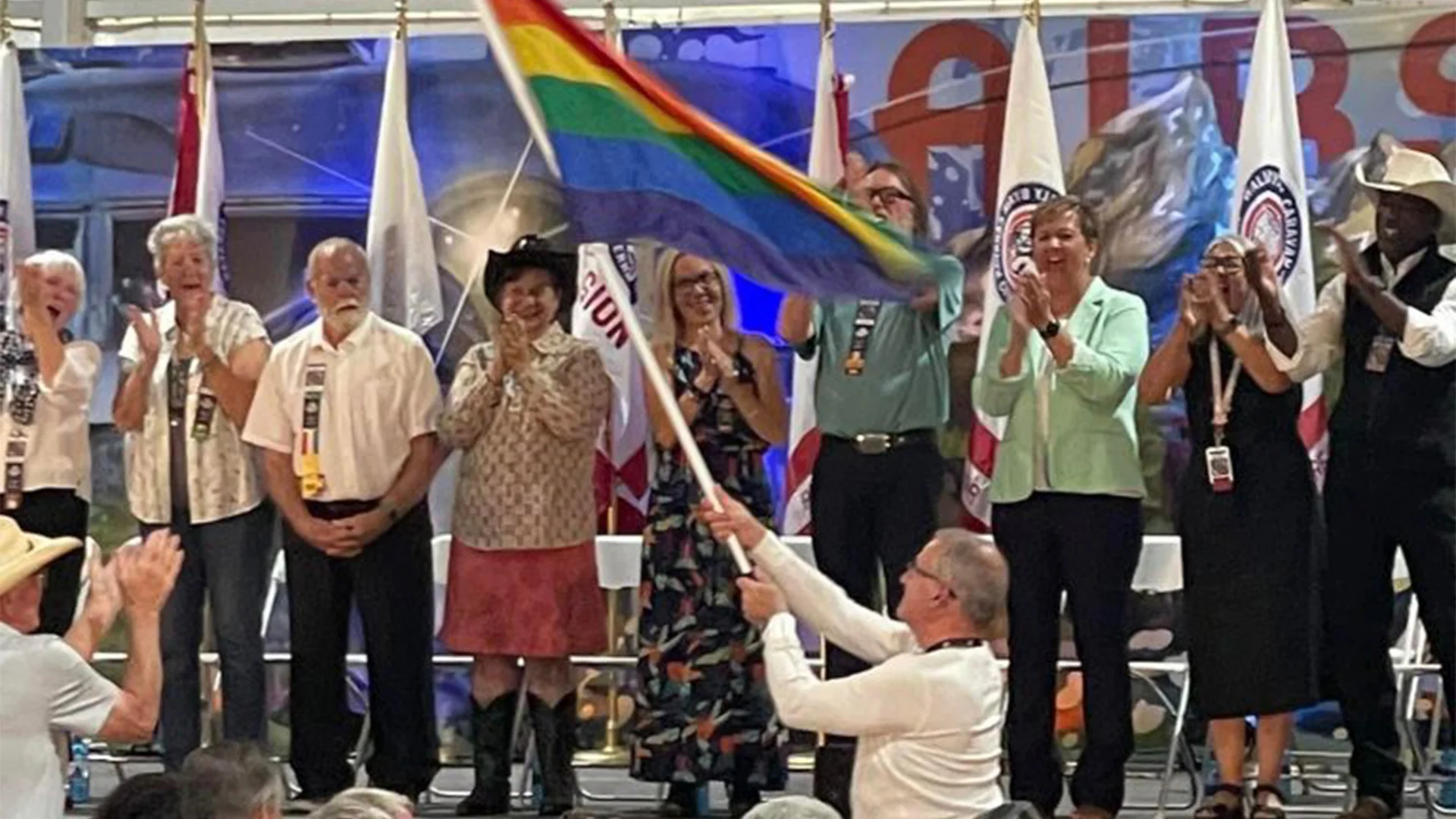 A person waves a rainbow flag on stage, while a lineup of people claps. Flags are displayed in the background.