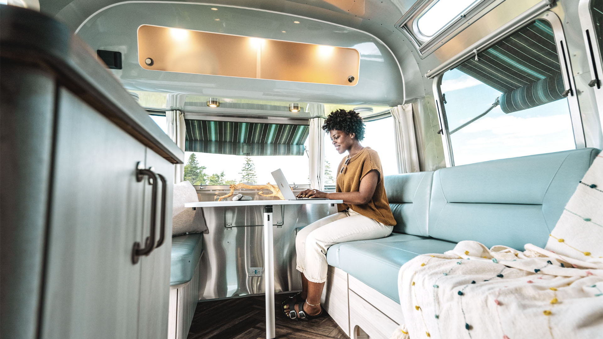 A woman sits on a light blue cushioned bench inside a modern RV, working on a laptop at a table. Sunlight streams in through large windows with striped awnings and scenic views outside.