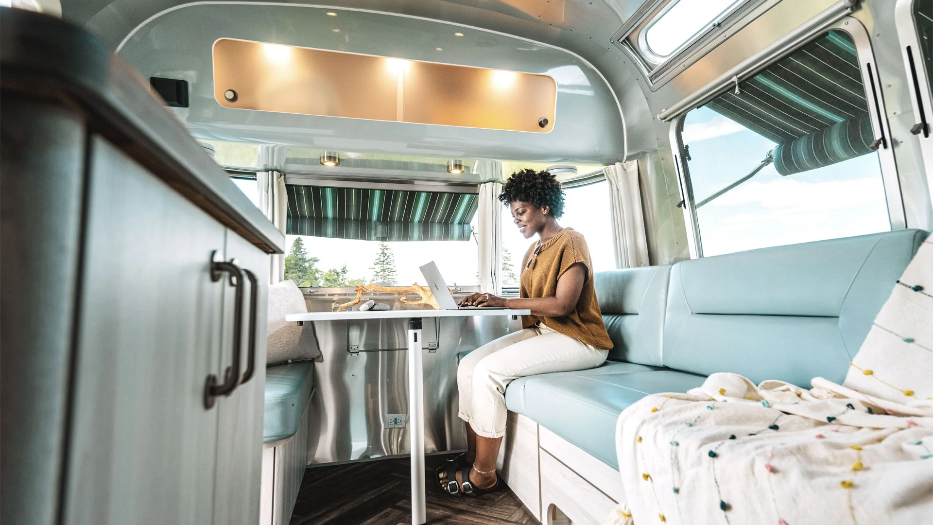 A woman sits on a light blue cushioned bench inside a modern RV, working on a laptop at a table. Sunlight streams in through large windows with striped awnings and scenic views outside.