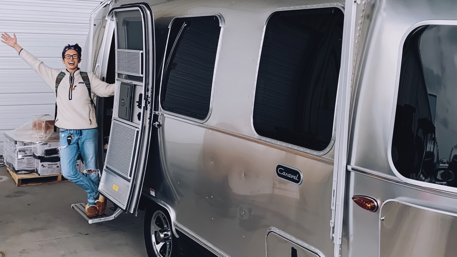 Person joyfully standing at the open door of a shiny silver Airstream trailer, wearing a backpack and casual attire.