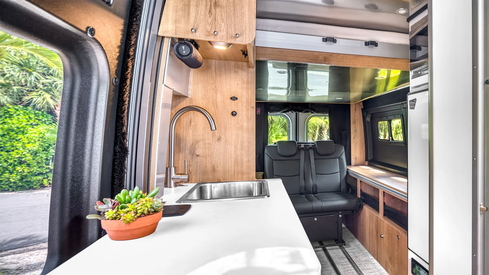 Modern camper van interior with a sleek kitchen, sink, wooden cabinets, black seating, and a small potted plant on the counter.