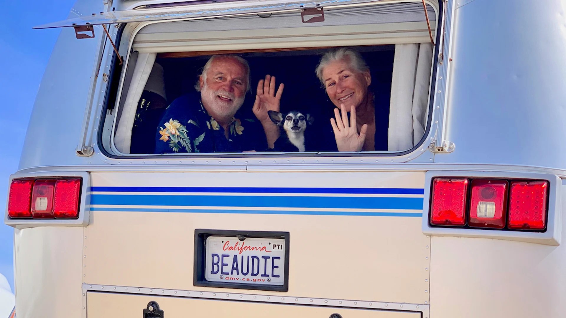 A smiling older man and woman wave from the open window of a white camper van with a small black and white dog between them. The license plate reads BEAUDIE and the sky is clear and blue.