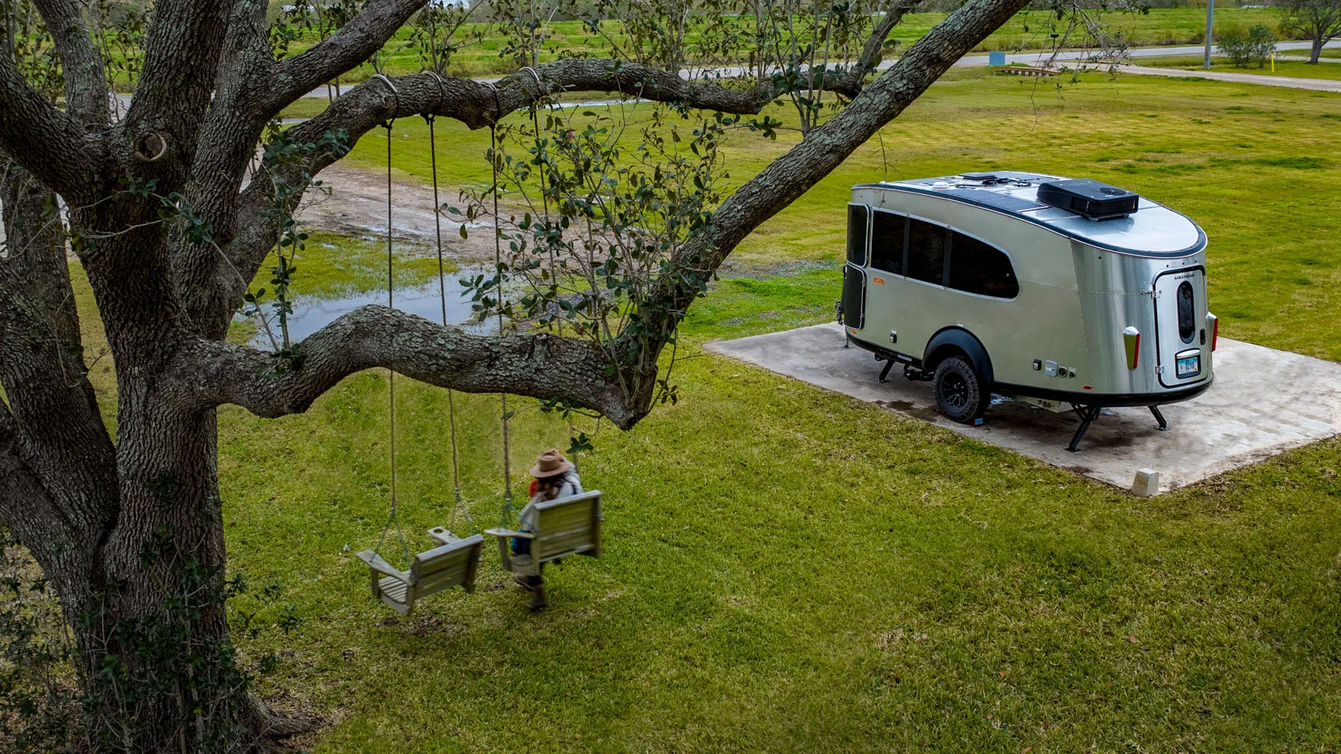 A person wearing a hat sits on a wooden swing hanging from a large tree, near a modern silver camper trailer parked on a grassy field.