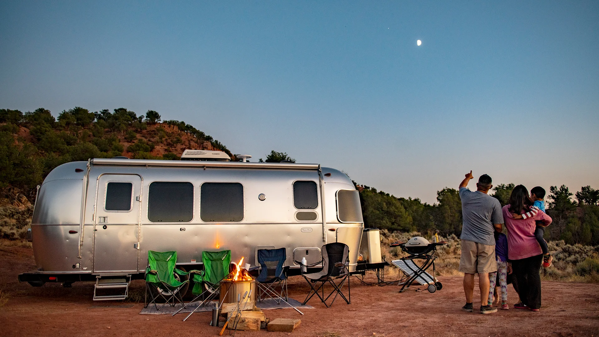 A family stands near a campfire and colorful chairs outside a silver Airstream trailer at dusk, looking at the sky. Trees and hills surround the campsite under a clear evening sky with a visible moon.
