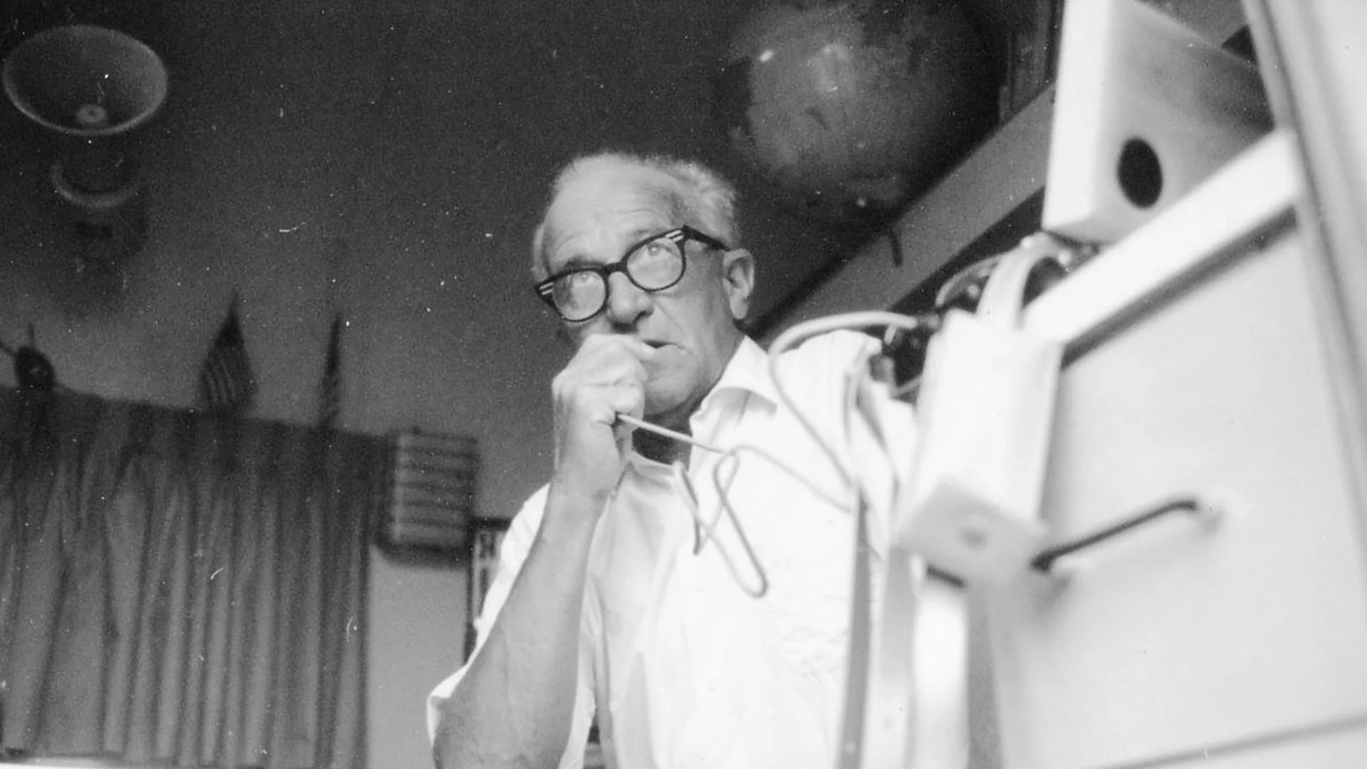 An older man with glasses holds a microphone to his mouth, appearing to speak. He stands indoors near equipment, with a globe and an American flag visible in the background.