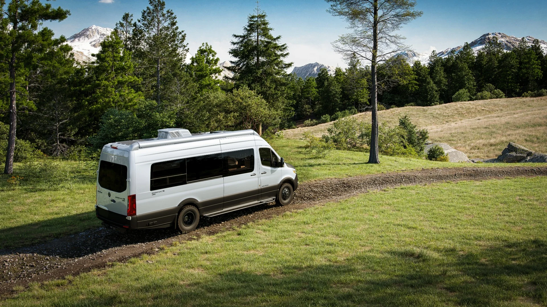 A white camper van drives on a gravel path through a scenic landscape with lush green trees and snow-capped mountains in the background.