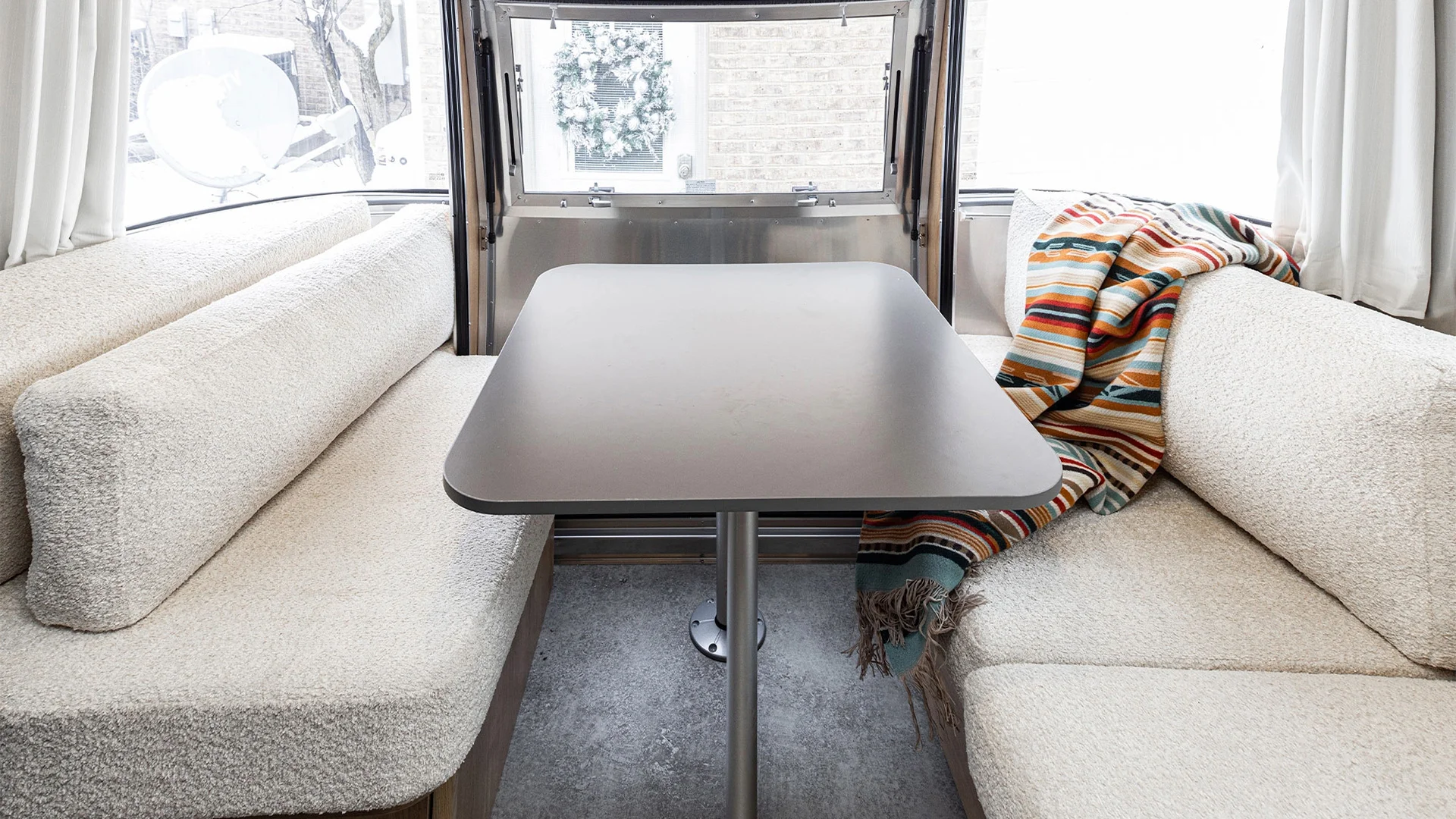Interior of a cozy RV dining area with a central table, cushioned benches, and a colorful striped blanket on the right bench.
