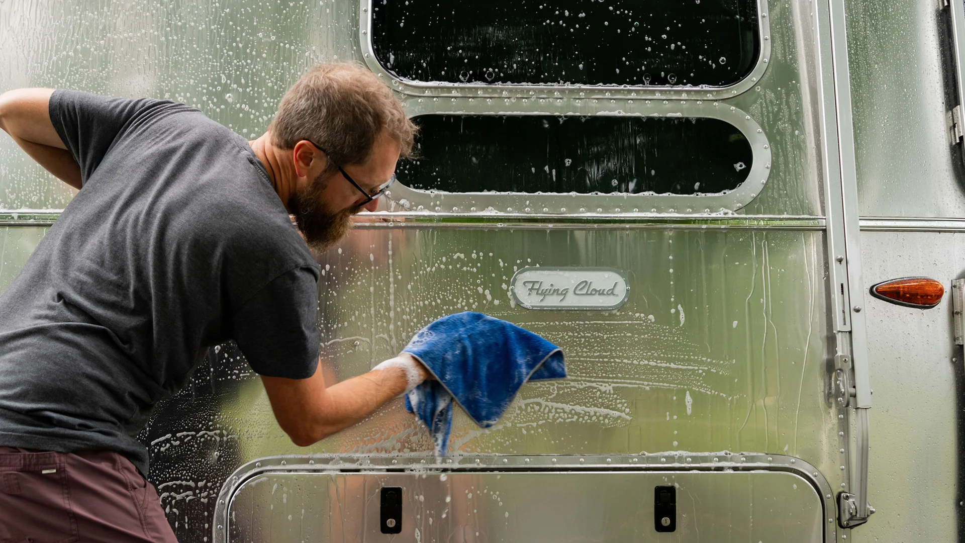 A person washes a shiny Airstream with a blue cloth and soapy water. The trailer has a "Flying Cloud" badge.