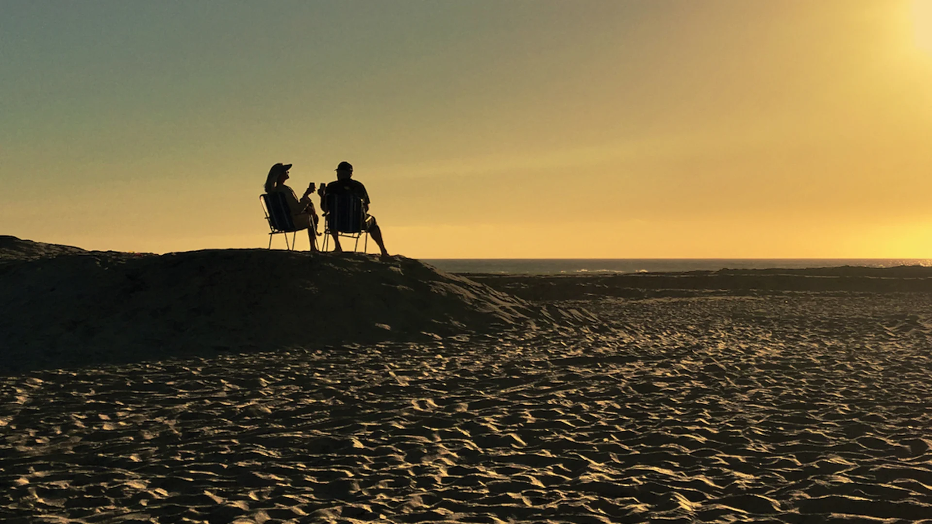 Two people sit in chairs on a sandy beach at sunset, silhouetted against the sky with the sun low on the horizon. The sea is calm in the background, creating a peaceful and relaxing atmosphere.