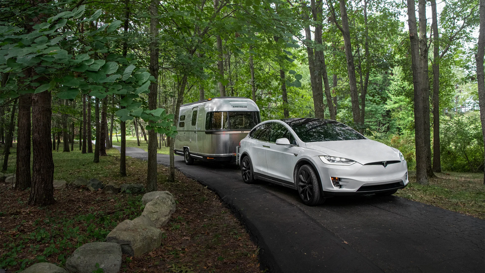 A white Tesla towing a silver Airstream trailer on a winding forest road, surrounded by lush green trees.