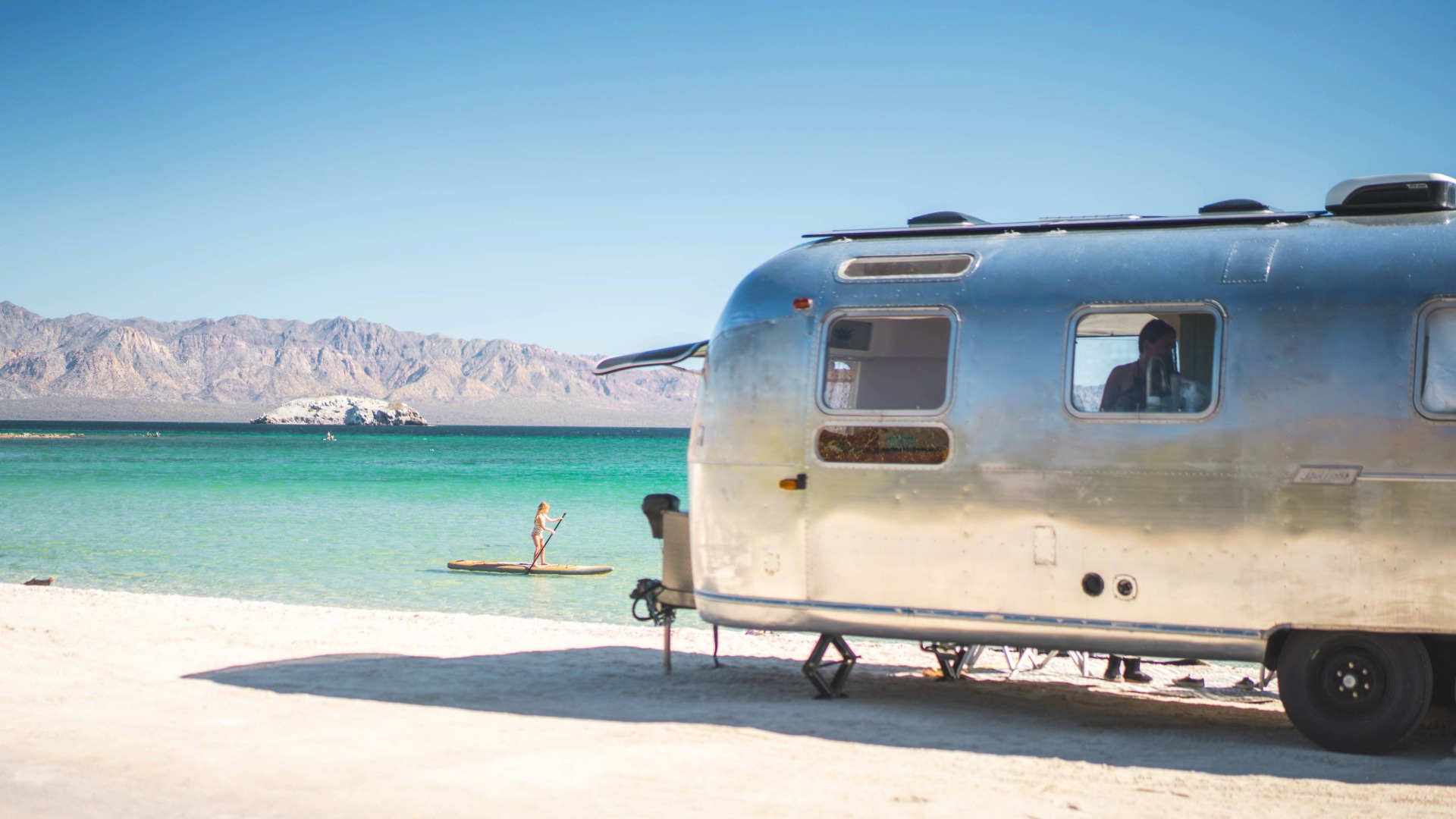 A shiny Airstream trailer sits on a sandy beach; a person paddleboarding on turquoise water with mountains in the background.
