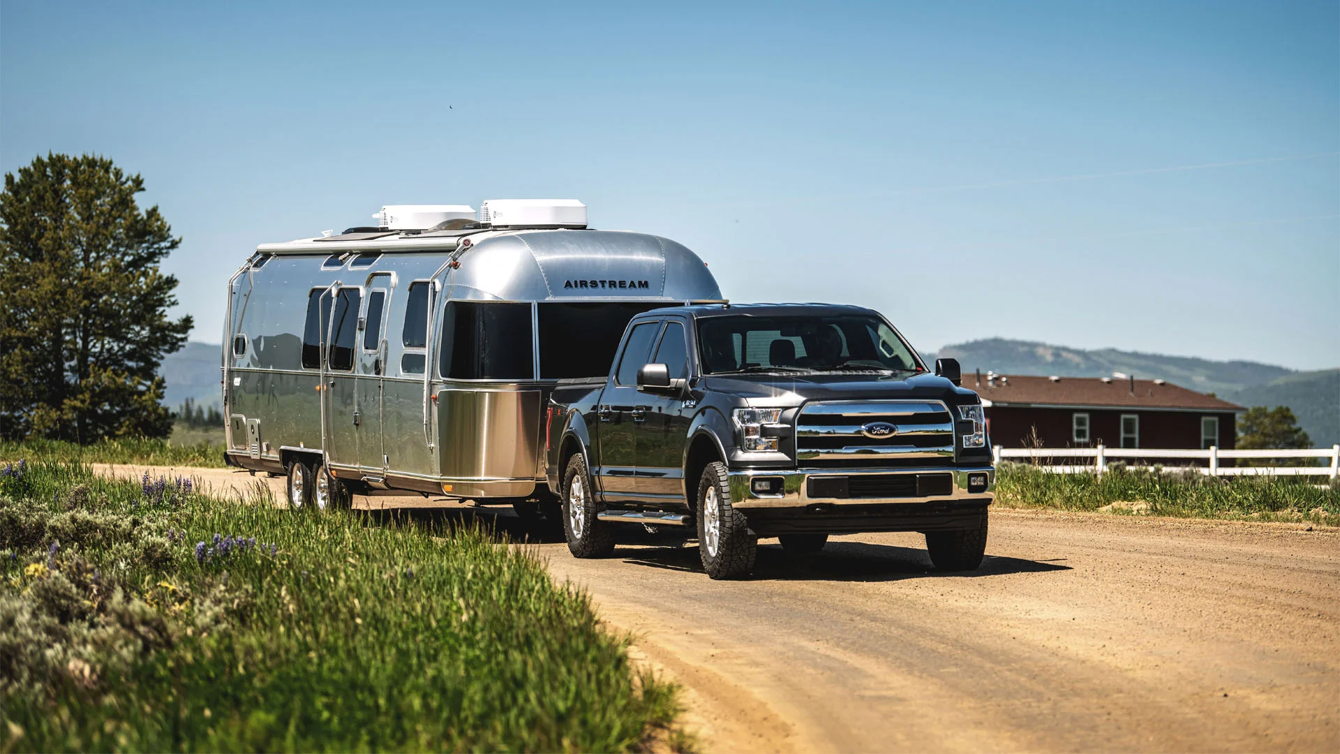 A black Ford pickup truck towing a silver Airstream travel trailer down a dirt road, with green grass, trees, and a house with mountains in the background under a clear blue sky.