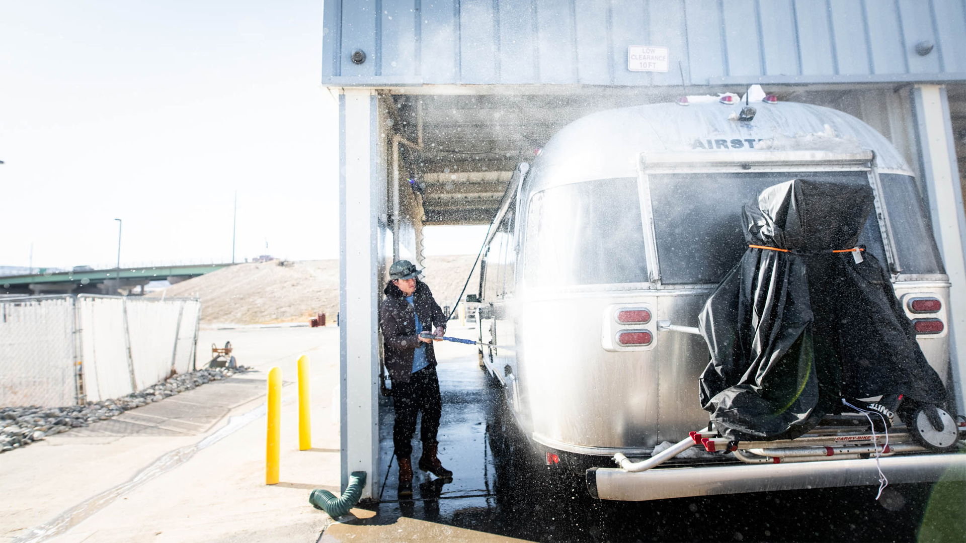 A person power washes a silver Airstream trailer at an outdoor car wash. Water spray fills the air as the trailer is cleaned under a metal roof, with sunlight brightening the scene.