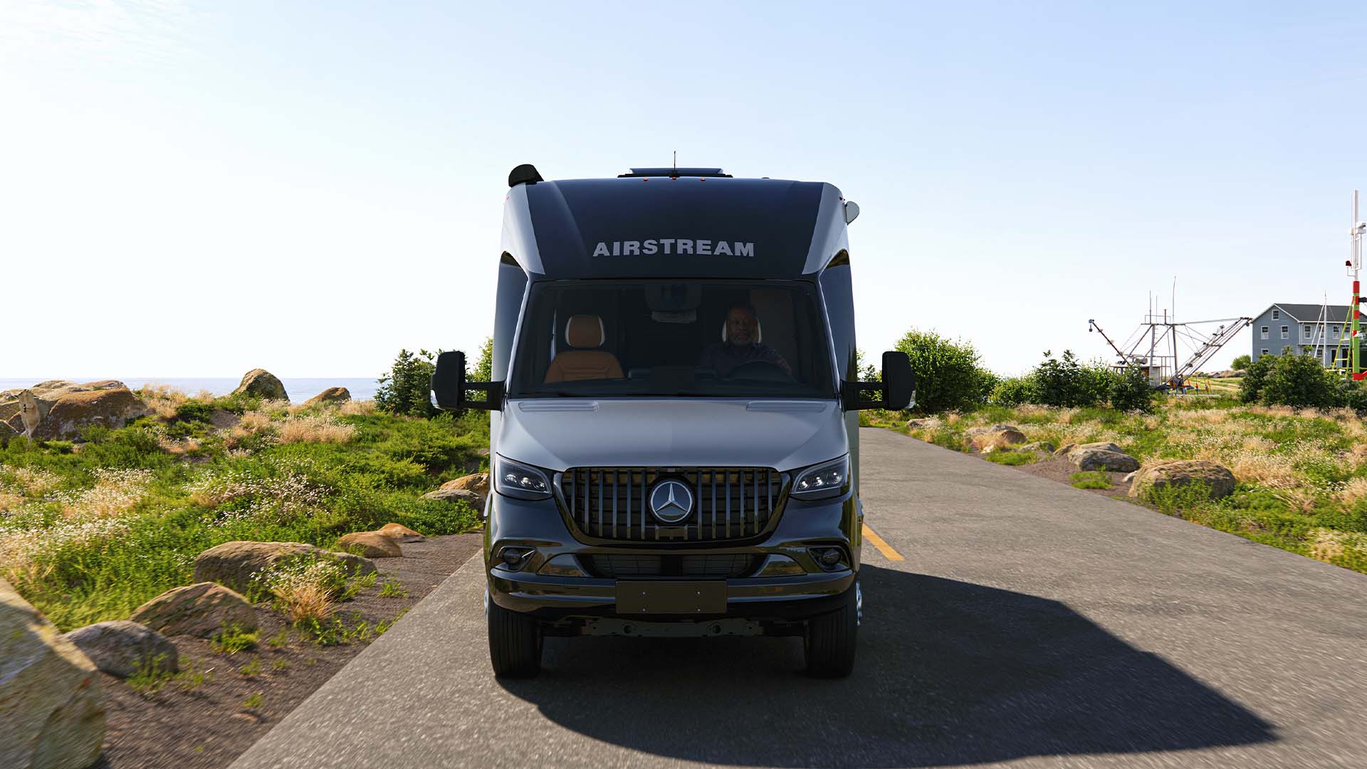 Front view of a silver Airstream van on a scenic rural road, surrounded by greenery and rocky landscape under a clear blue sky.