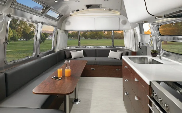 Modern RV interior with a gray seating area, wooden table, kitchenette with stainless sink, and large windows showing a park view.