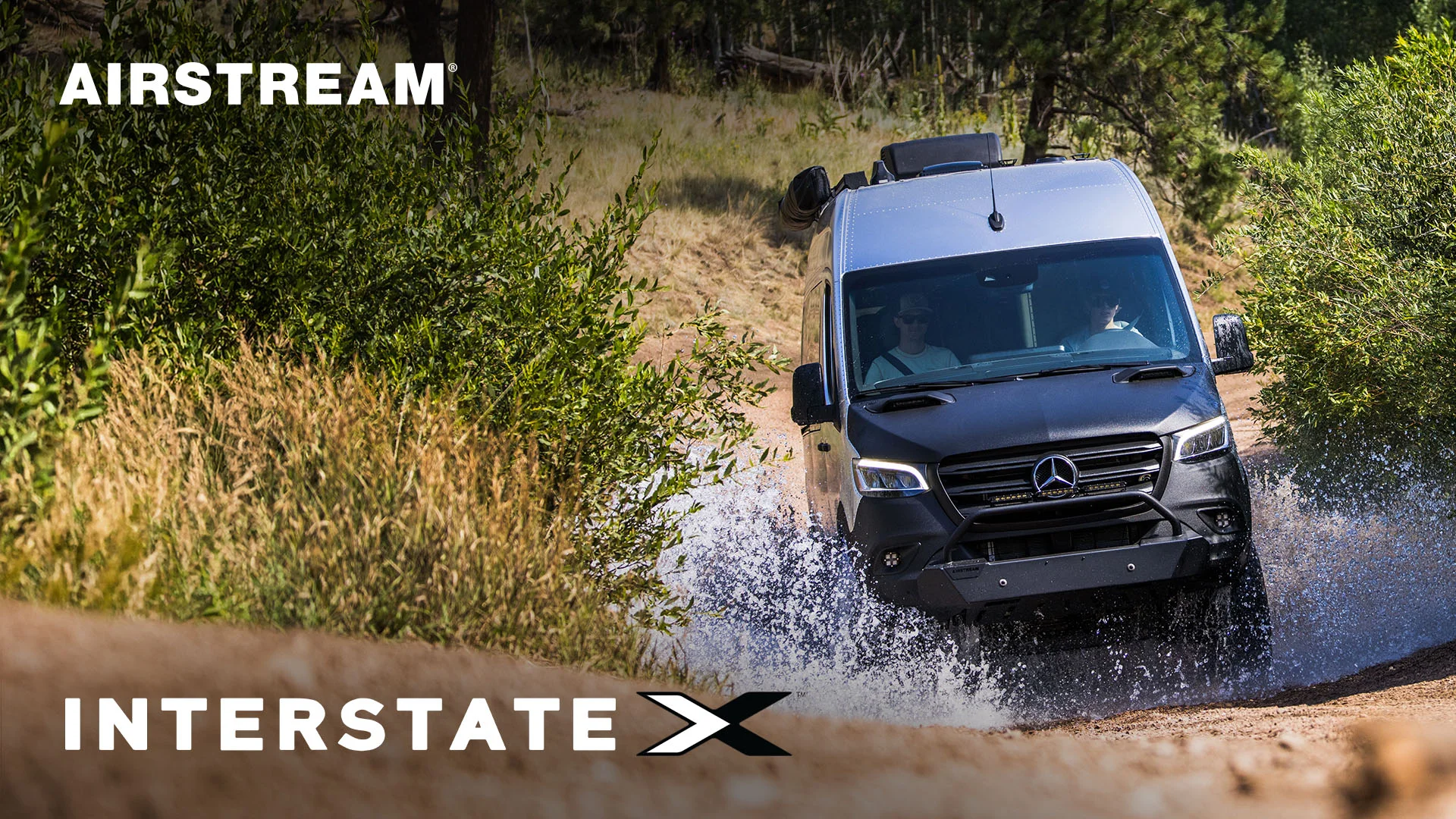 Airstream Interstate X van driving through water on a dirt trail, surrounded by greenery, under sunny sky.