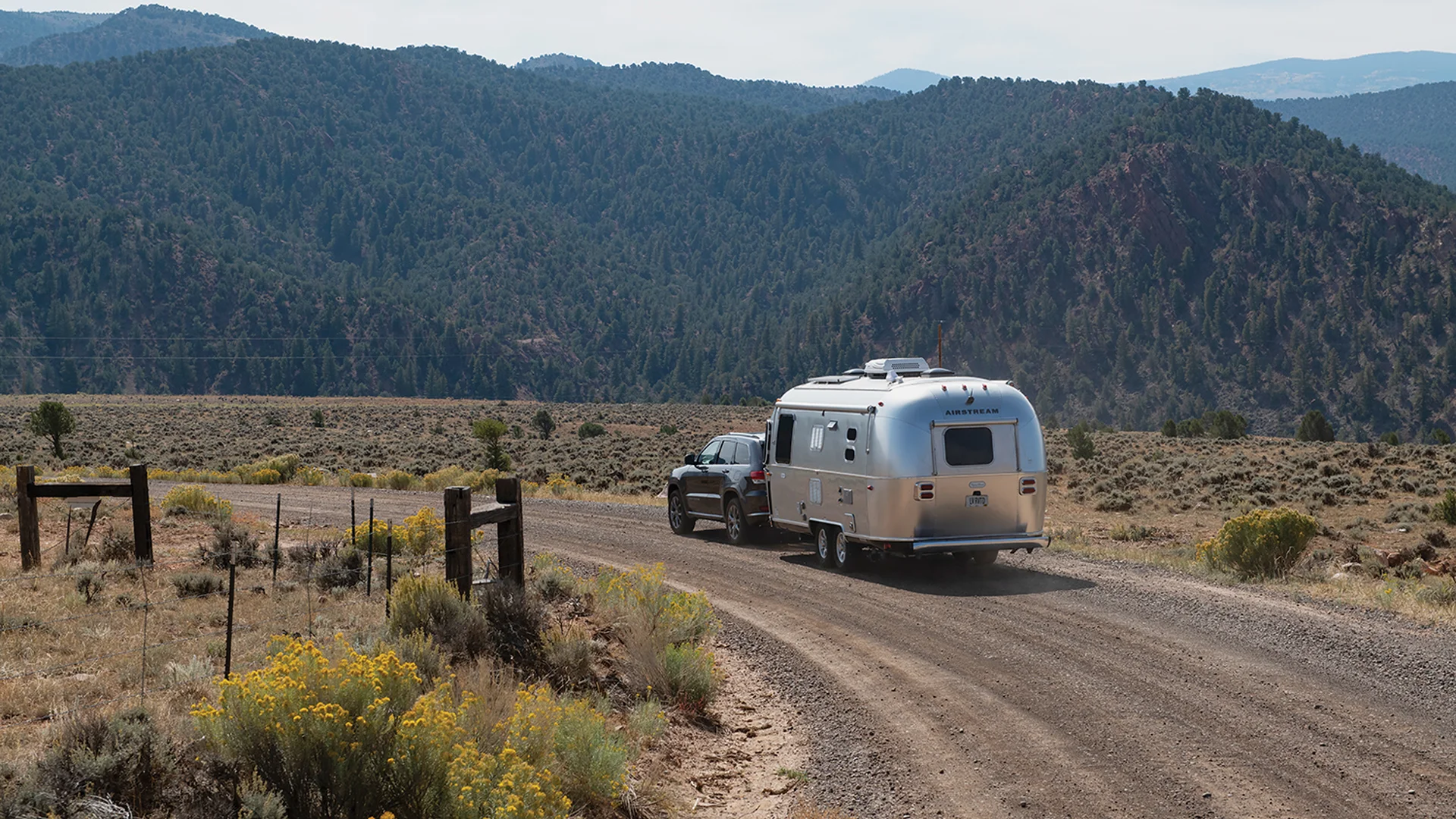 A silver Airstream trailer is being towed by a dark SUV along a winding dirt road, surrounded by dry grass and shrubs, with forested mountains rising in the background under a hazy sky.
