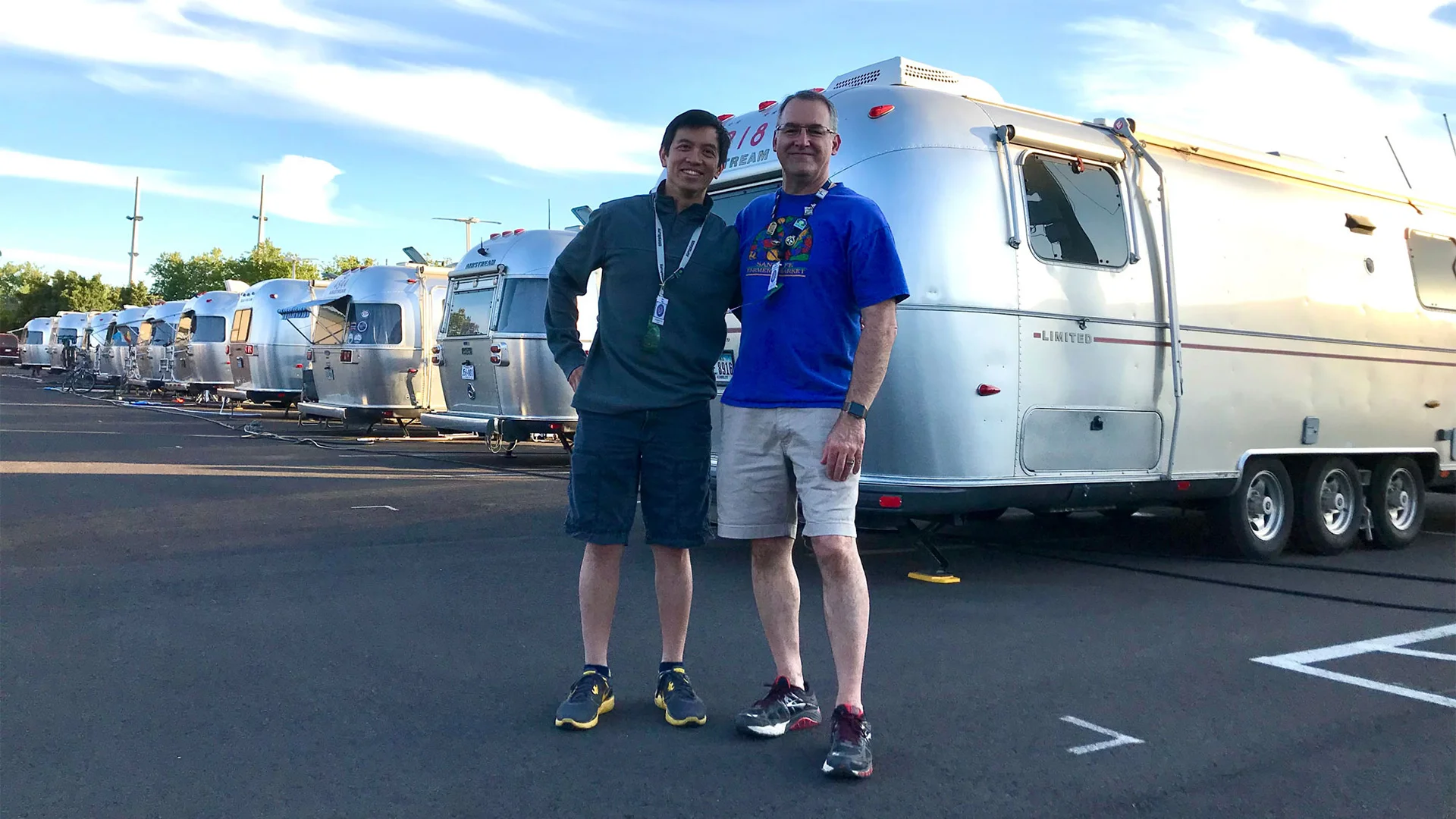 Two people standing arm in arm in front of a row of silver Airstream trailers in a parking area under a partly cloudy sky.