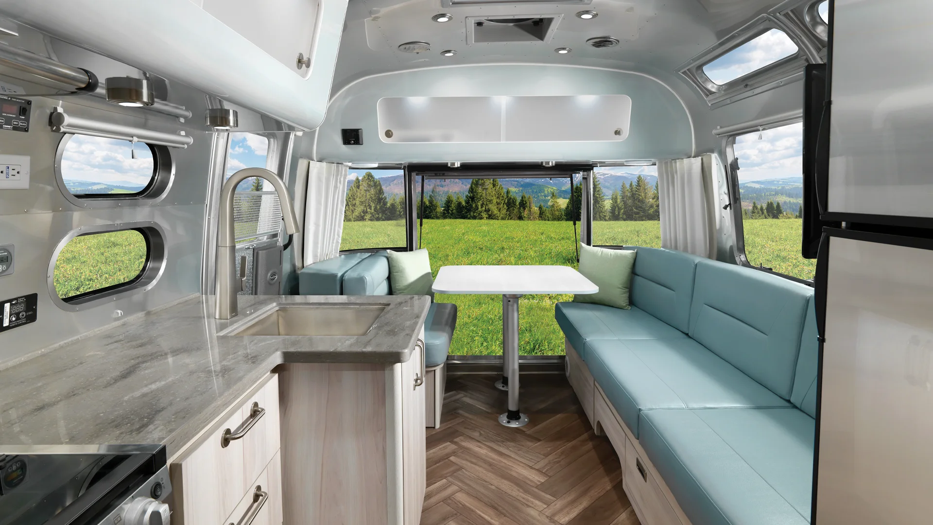 Interior of a modern camper van with light blue seating, a small table, kitchen area, and large windows overlooking a green field and trees under a blue sky.